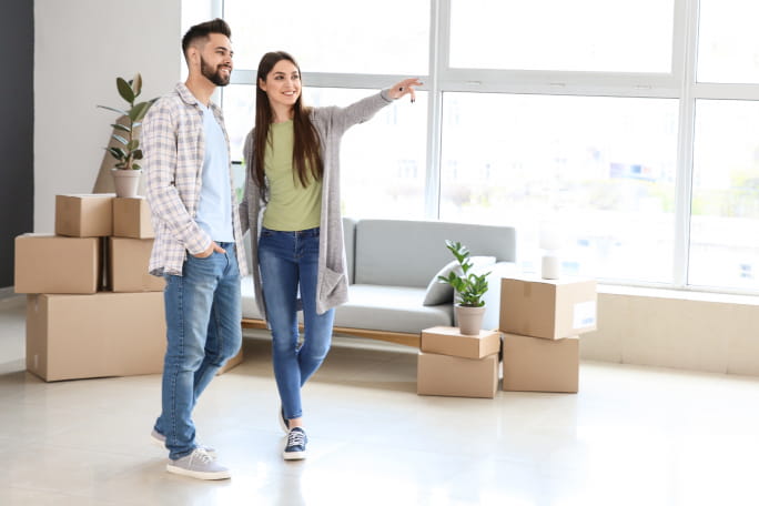 Homem e mulher jovens numa sala de uma casa com caixas ao fundo.