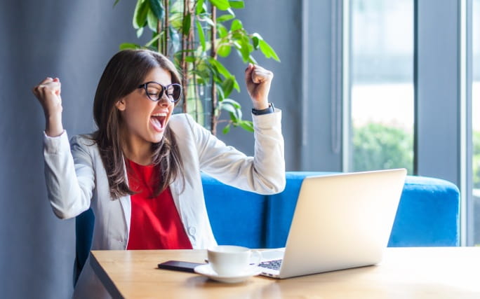 Jovem mulher, sentada em frente a um portátil, festeja os objetivos financeiros que alcançou.