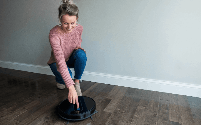 Uma jovem feliz em casa liga o aspirador robot para limpar o chão