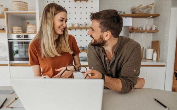 Um casal jovem numa cozinha, conversa animadamente  de frente para um laptop aberto. 