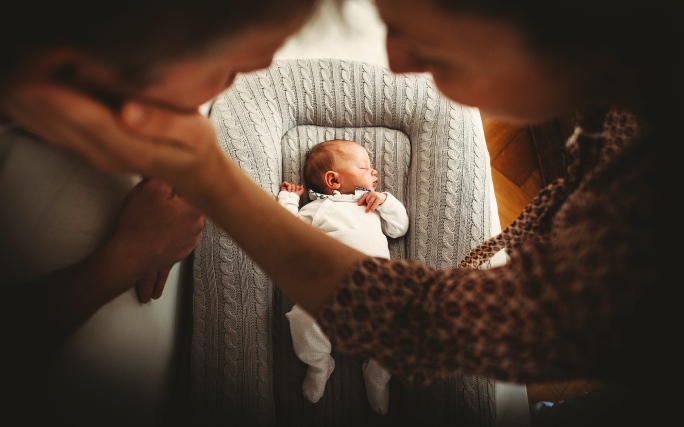 Recém‑nascido a dormir num ninho acolchoado, com os pais inclinados sobre ele num momento terno.