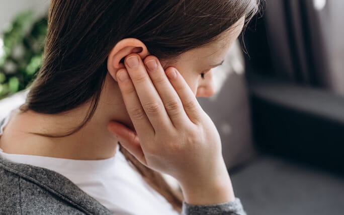 Mulher dentro de casa com ar de sofrimento aperta a orelha para tentar atenuar a dor de ouvido