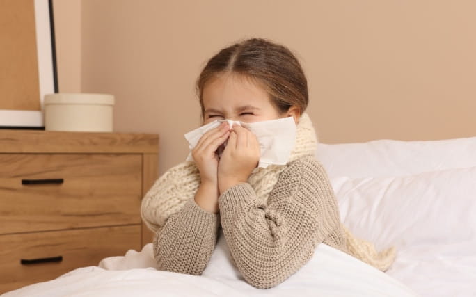 Uma menina sentada numa cama assoa o nariz com força.