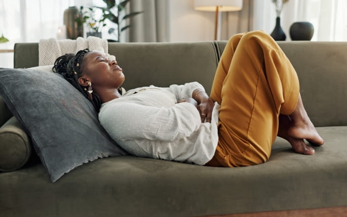 Mulher com expressão de desconforto, deitada num sofá em casa agarrada à barriga por sofrer de dores de estômago.