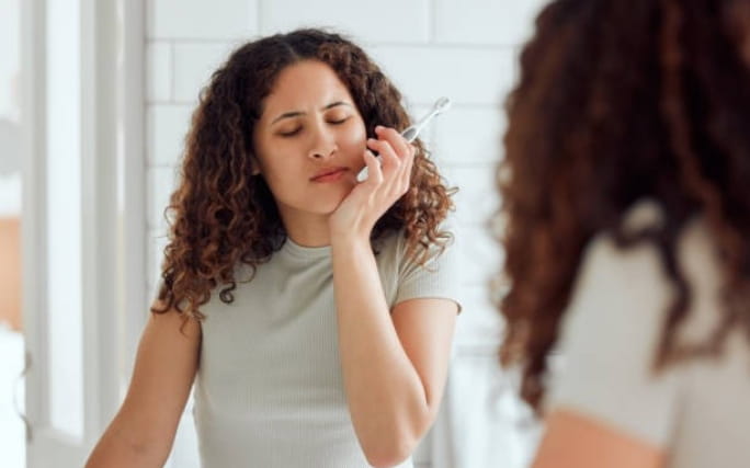 Mulher jovem frente a um espelho de casa de banho mostra sinais de dor de dentes por sensibilidade dentária ao escovar os dentes.