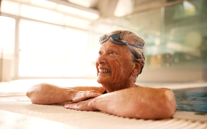 Mulher idosa a sorrir dentro de uma piscina interior com óculos de natação.