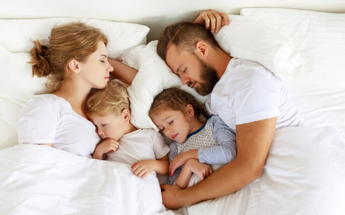 Família de pai, mãe e dois filhos deitados a dormir na mesma cama.