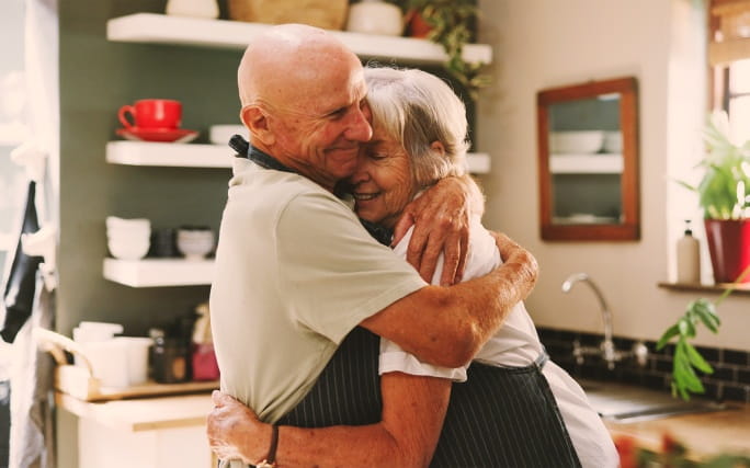 Casal idoso a abraçar-se carinhosamente na cozinha.