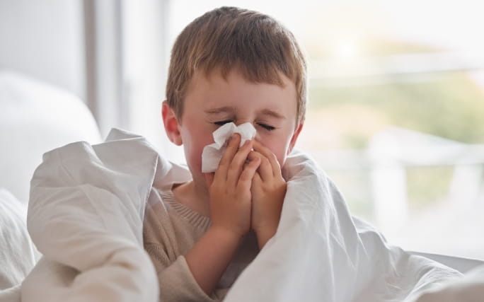 Menino numa cama com ar adoentado, assoa o nariz devido à sinusite infantil.