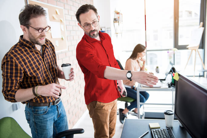 Dois homens no escritório a conversarem e a apontarem para um computador.