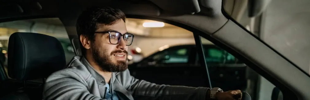 Homem jovem de barba e óculos no interior de um carro estacionado numa garagem.